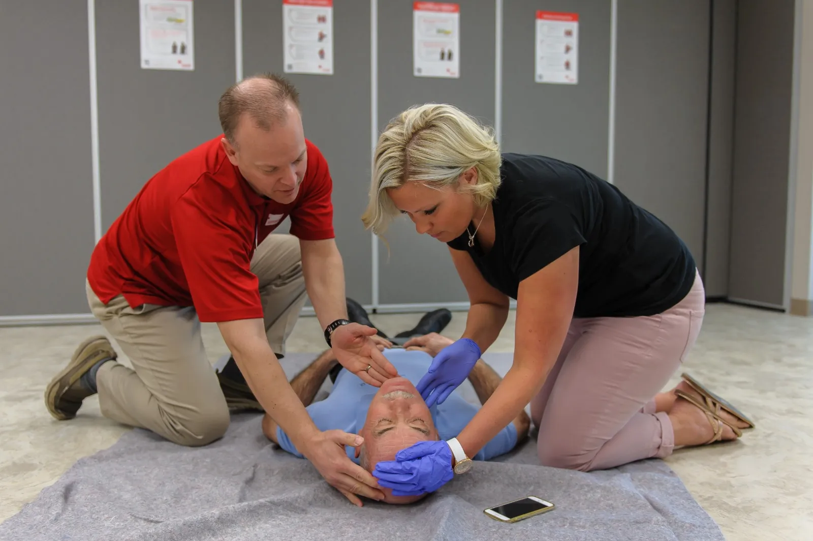 First aid instructor guiding a student as she opens the airway of a person lying on the floor during a first aid class